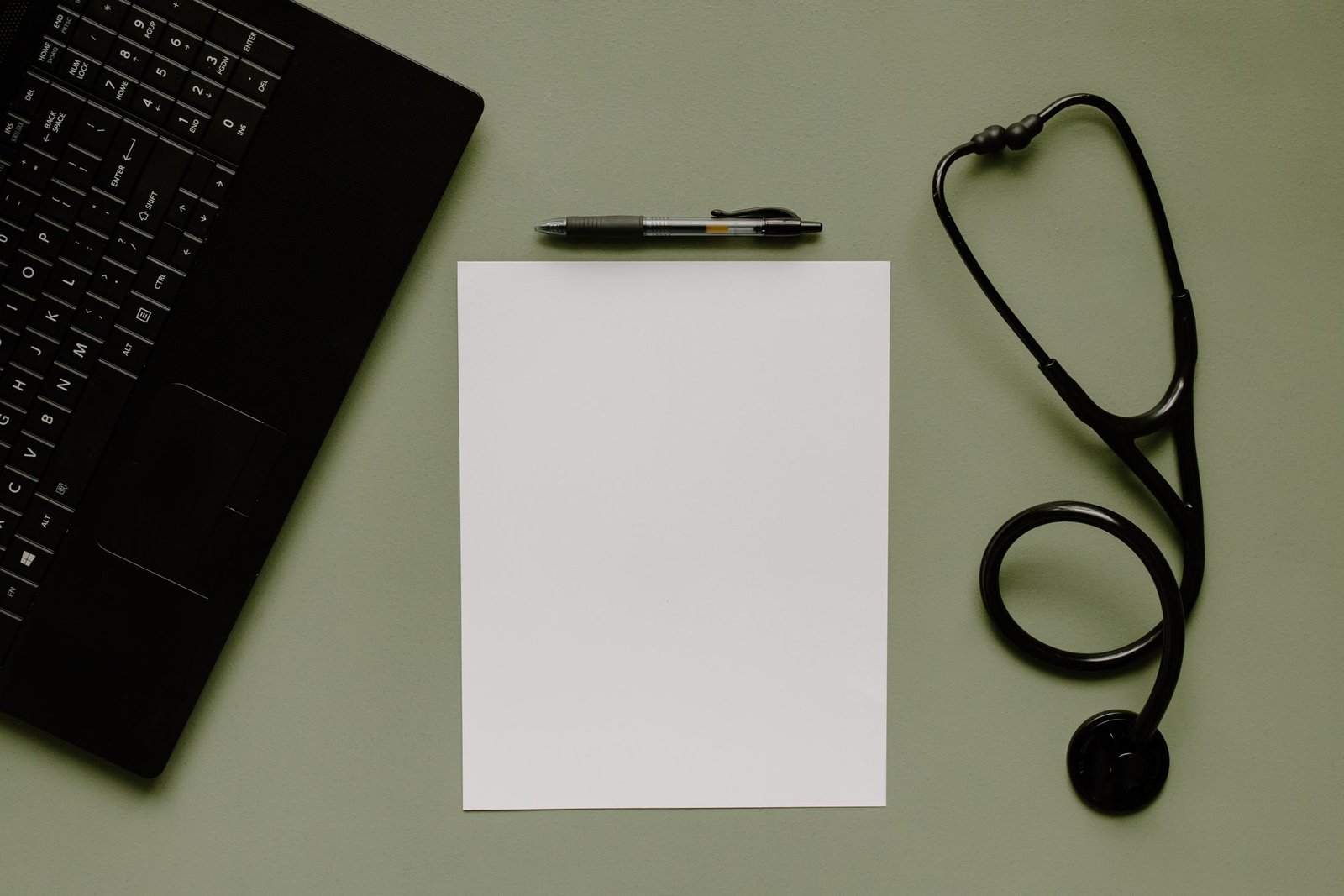 Medical documentation workspace with a laptop, blank paper, pen and stethoscope.